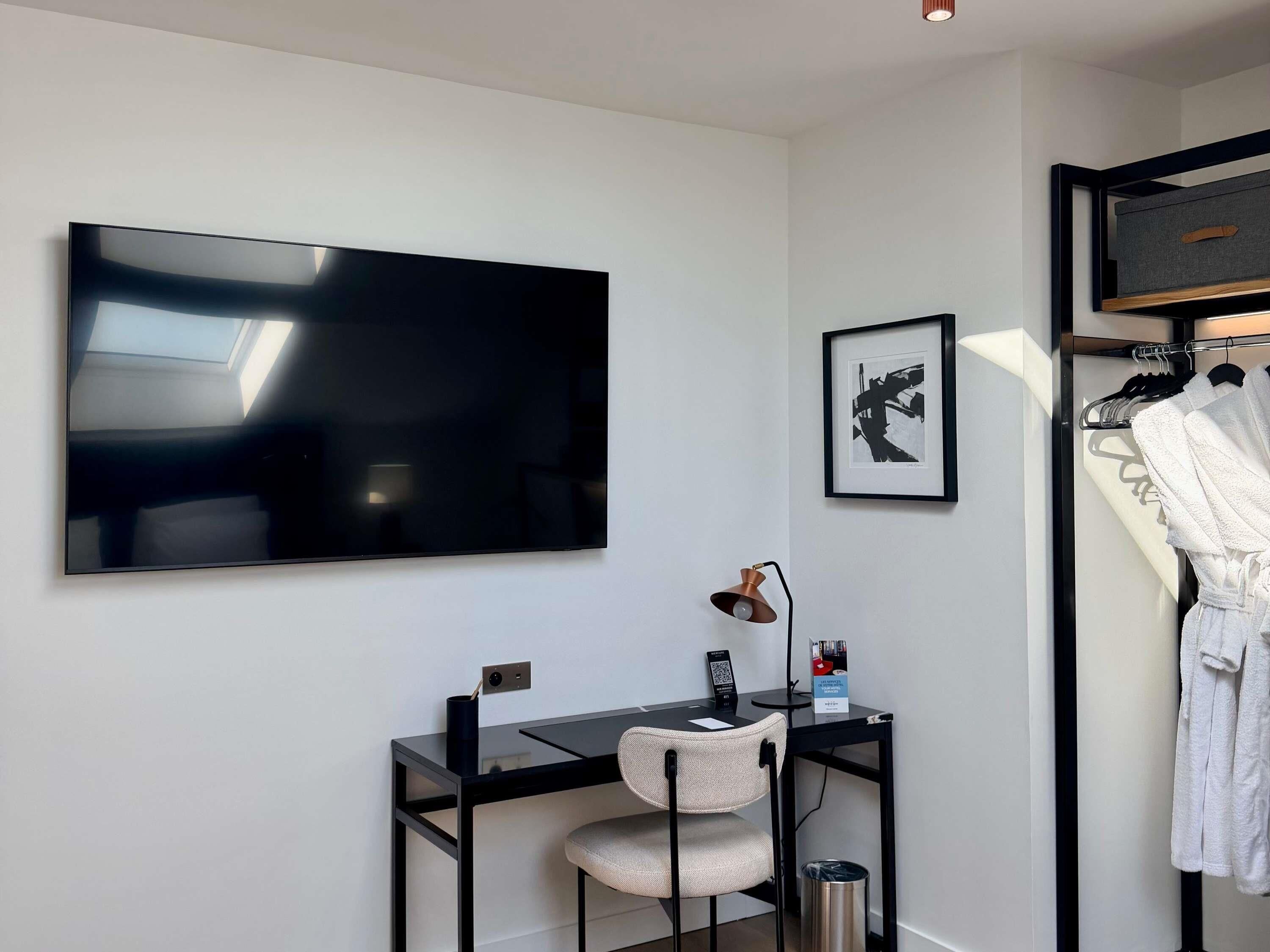 A modern bedroom with a large flat-screen TV, a desk with a chair, a framed picture, and a clothing rack with robes.
