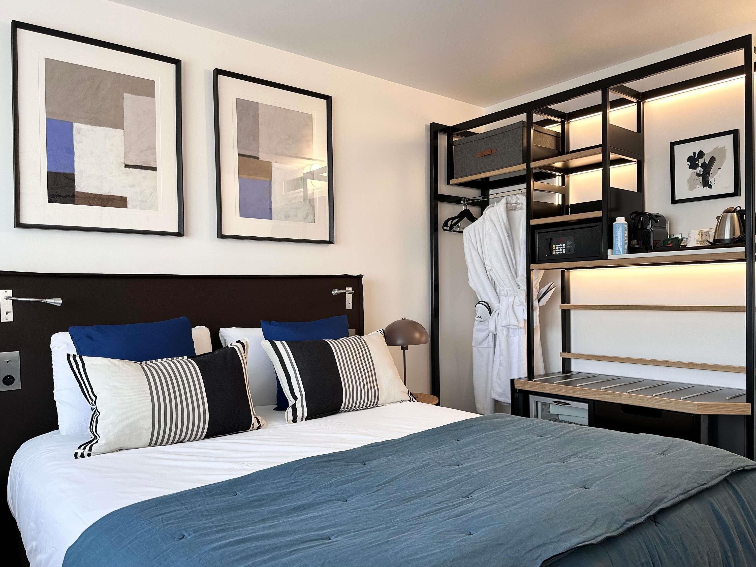 Una habitación de hotel con cama, mesita de noche, bata colgada, estante con artículos de aseo y dos cuadros abstractos enmarcados en la pared.