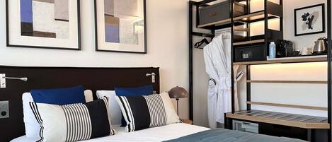 A hotel room with a bed, a nightstand, a robe hanging, a shelf with toiletries, and two framed abstract paintings on the wall.