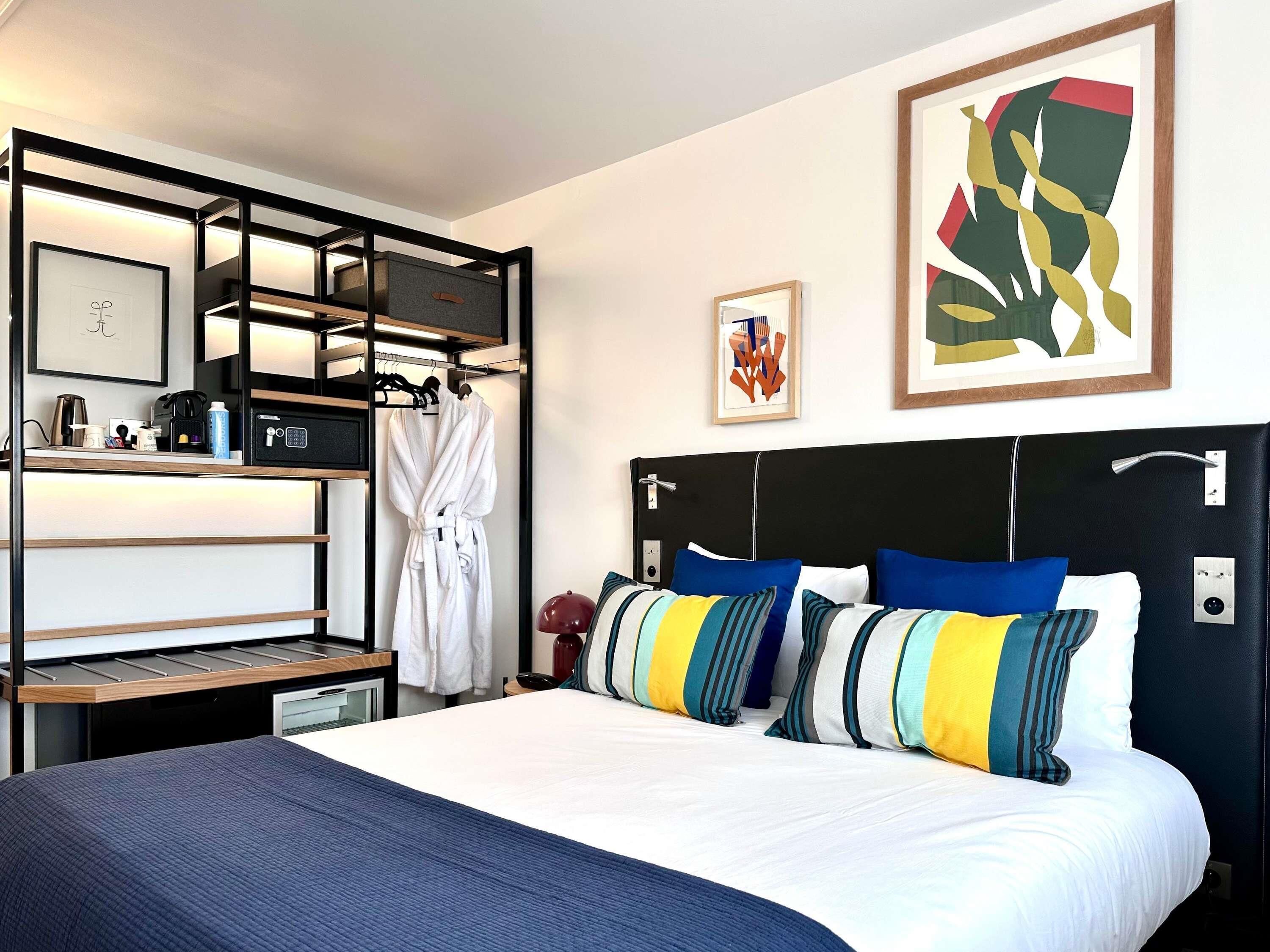 A hotel room with a bed, a bedside table, a built-in shelf, a robe, and artwork on the wall.