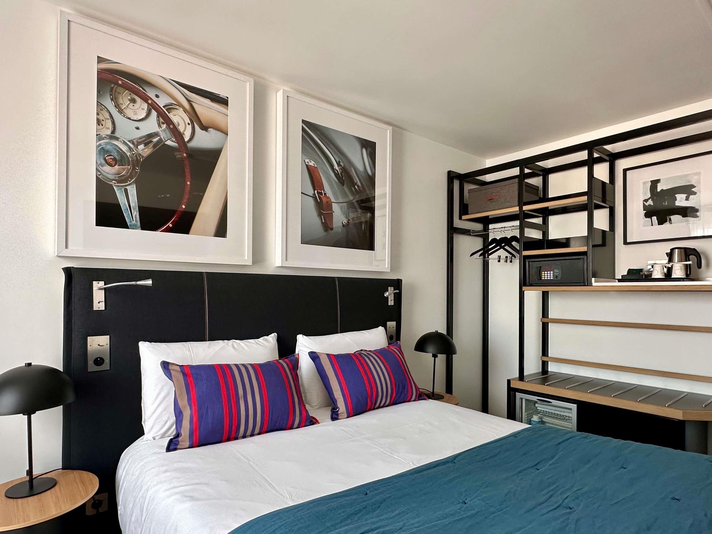 Una habitación de hotel con una cama, dos cuadros enmarcados, una mesita de noche, una lámpara, un estante con objetos y un perchero en la pared.