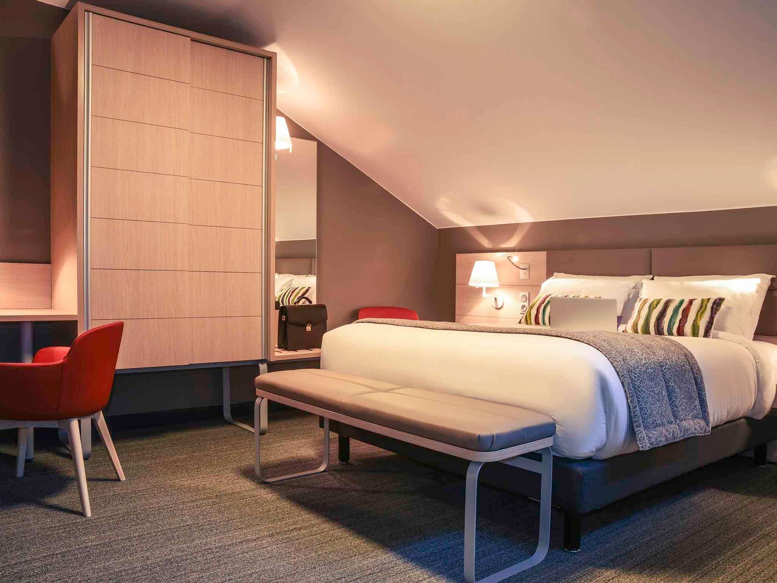 Ein Hotelzimmer mit Bett, Nachttisch, Lampe, einem roten Sessel und einem Kleiderschrank.