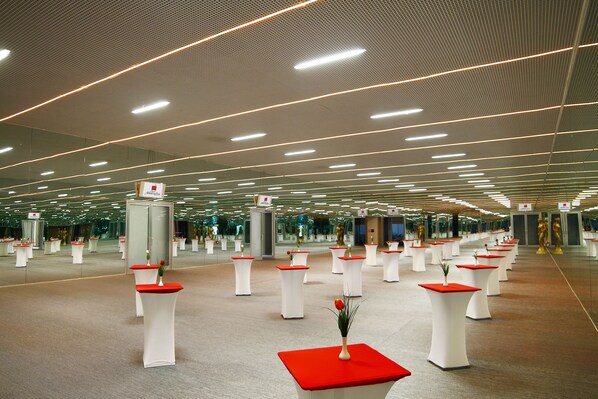 A spacious indoor area with multiple white tables, each topped with a red surface and a small plant. The ceiling features linear lighting fixtures, and the floor is carpeted.