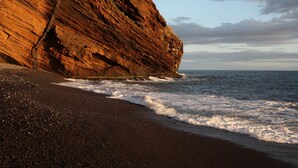 Litoral rochoso com um penhasco imponente e ondas quebrando em uma praia de pedras.