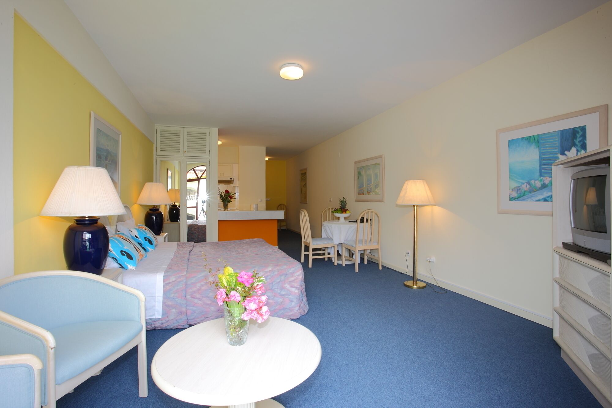 Une chambre d’hôtel avec un lit, deux chaises, une petite table décorée de fleurs, un téléviseur et, au fond, une kitchenette.