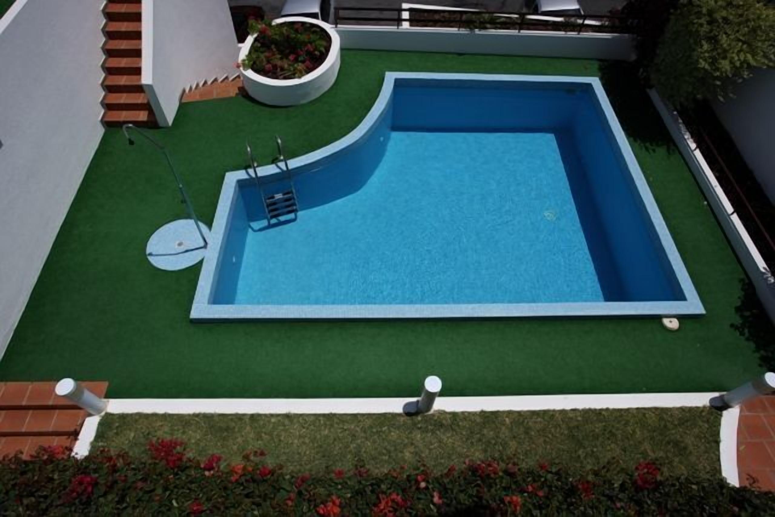 A rectangular swimming pool with a white border, surrounded by artificial green grass, and a small white planter with red flowers.