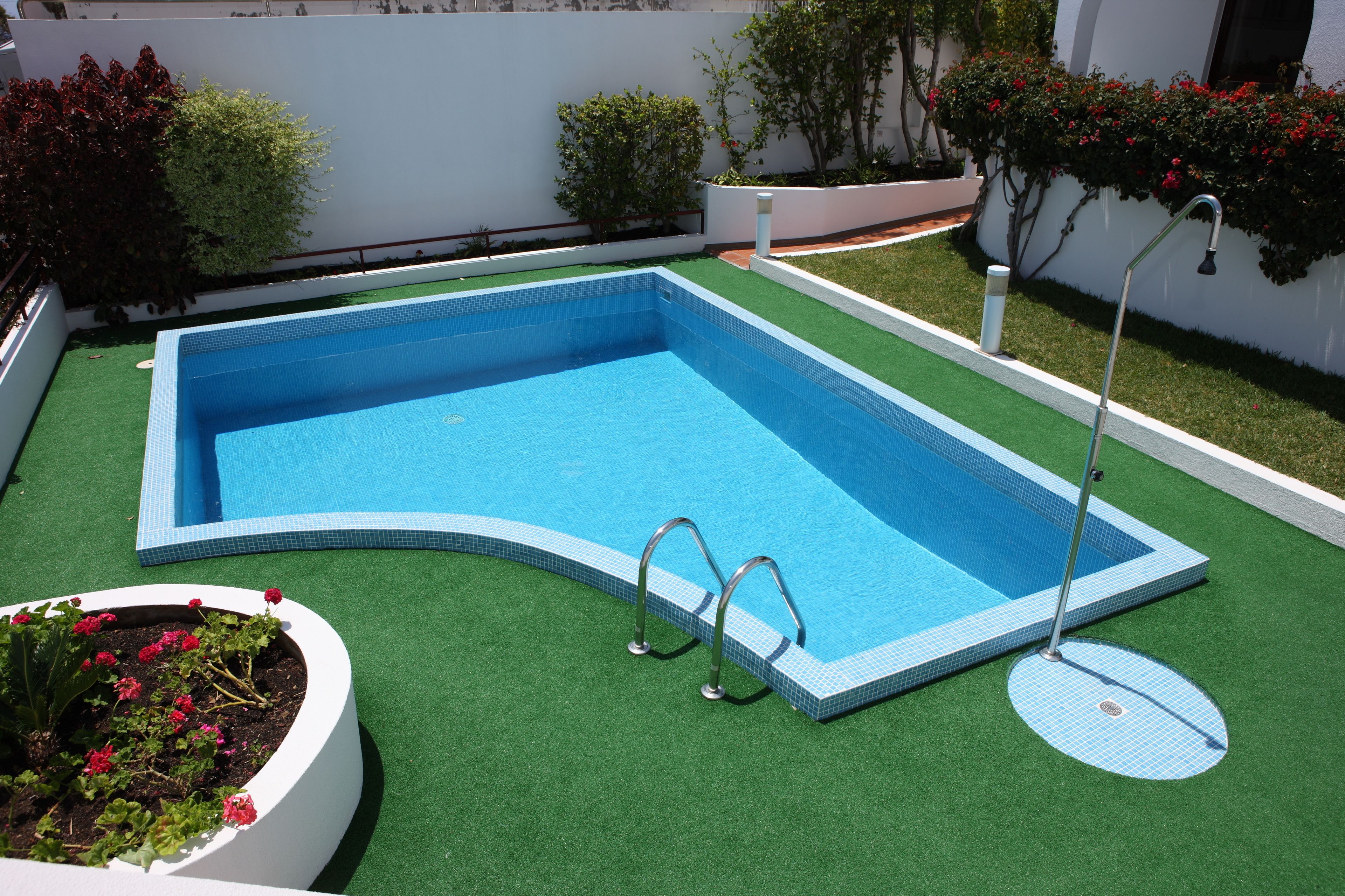 A rectangular swimming pool with a tiled edge, surrounded by artificial grass and bordered by white walls.