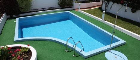 A rectangular swimming pool with a tiled edge, surrounded by artificial grass and bordered by white walls.