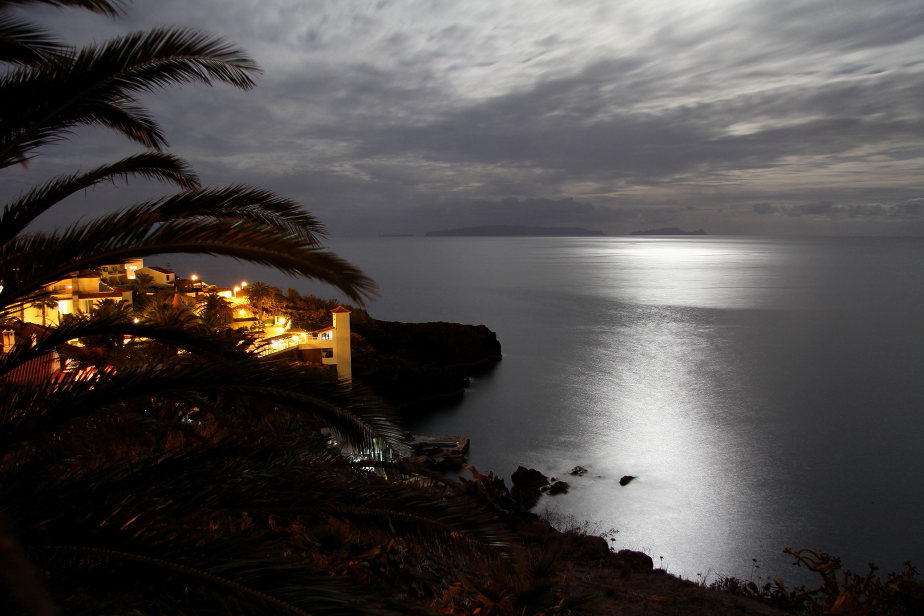 Une scène côtière avec un bâtiment illuminé par des lumières artificielles, surplombant une mer calme sous un ciel nuageux.