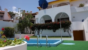Piscina com guarda-sol azul, rodeada por vegetação e um edifício branco com arcos.