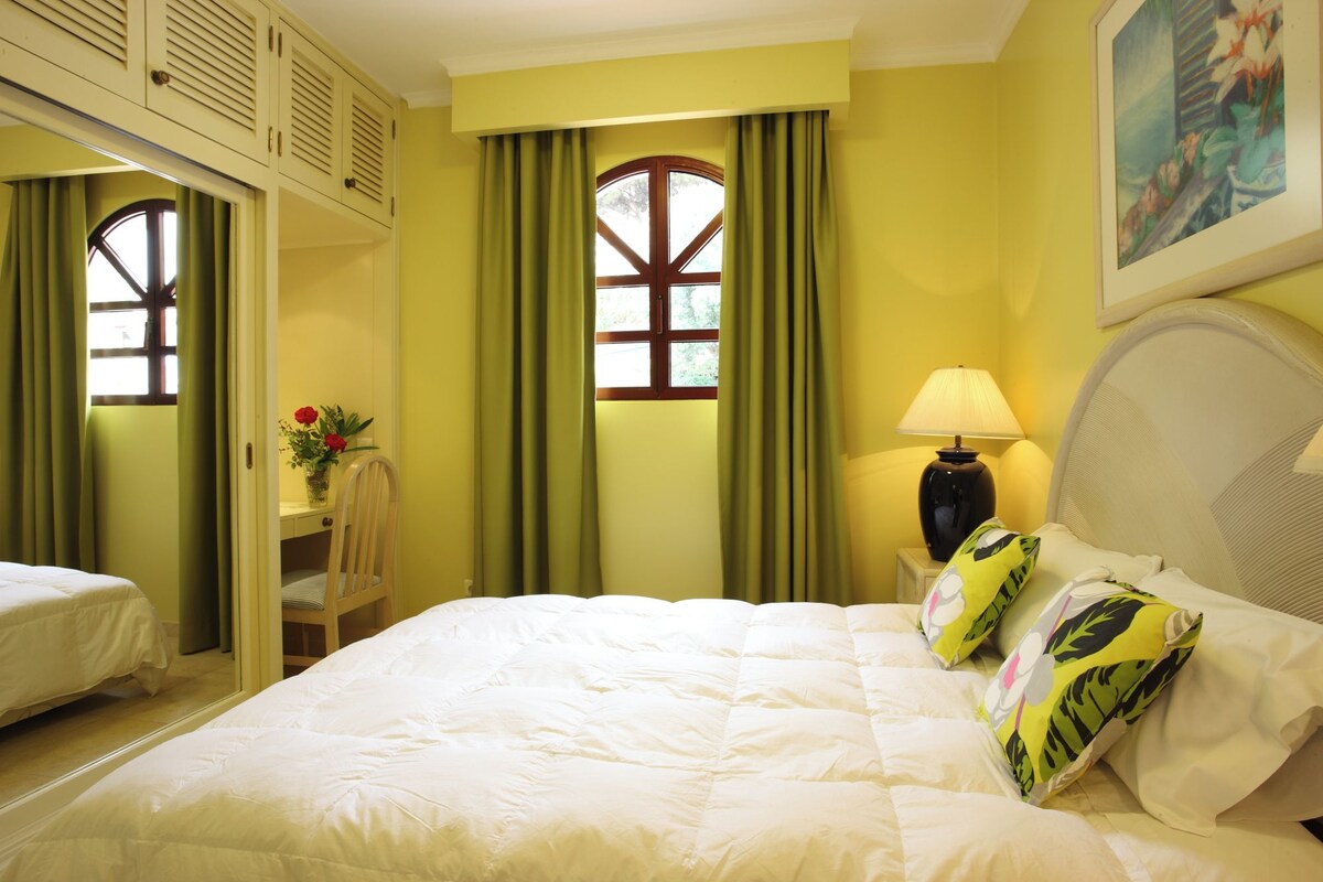 A hotel room with a large bed, a nightstand with a lamp, a chair, a desk, and a window with curtains.