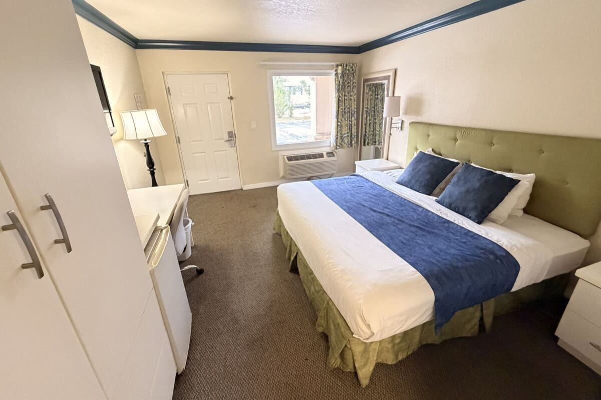 A hotel room with a bed, a desk, a chair, a window, and a door.
