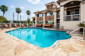A swimming pool with a stone deck, lounge chairs, and a building with balconies in the background.