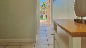 A hallway with a tiled floor, a painting on the wall, a lamp, and a view of a pool area. - Creekside Inn (Tavernier)