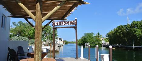 A wooden pergola with a sign reading "CREEKSIDE" at a dock with chairs and tables, overlooking a calm waterway and greenery.