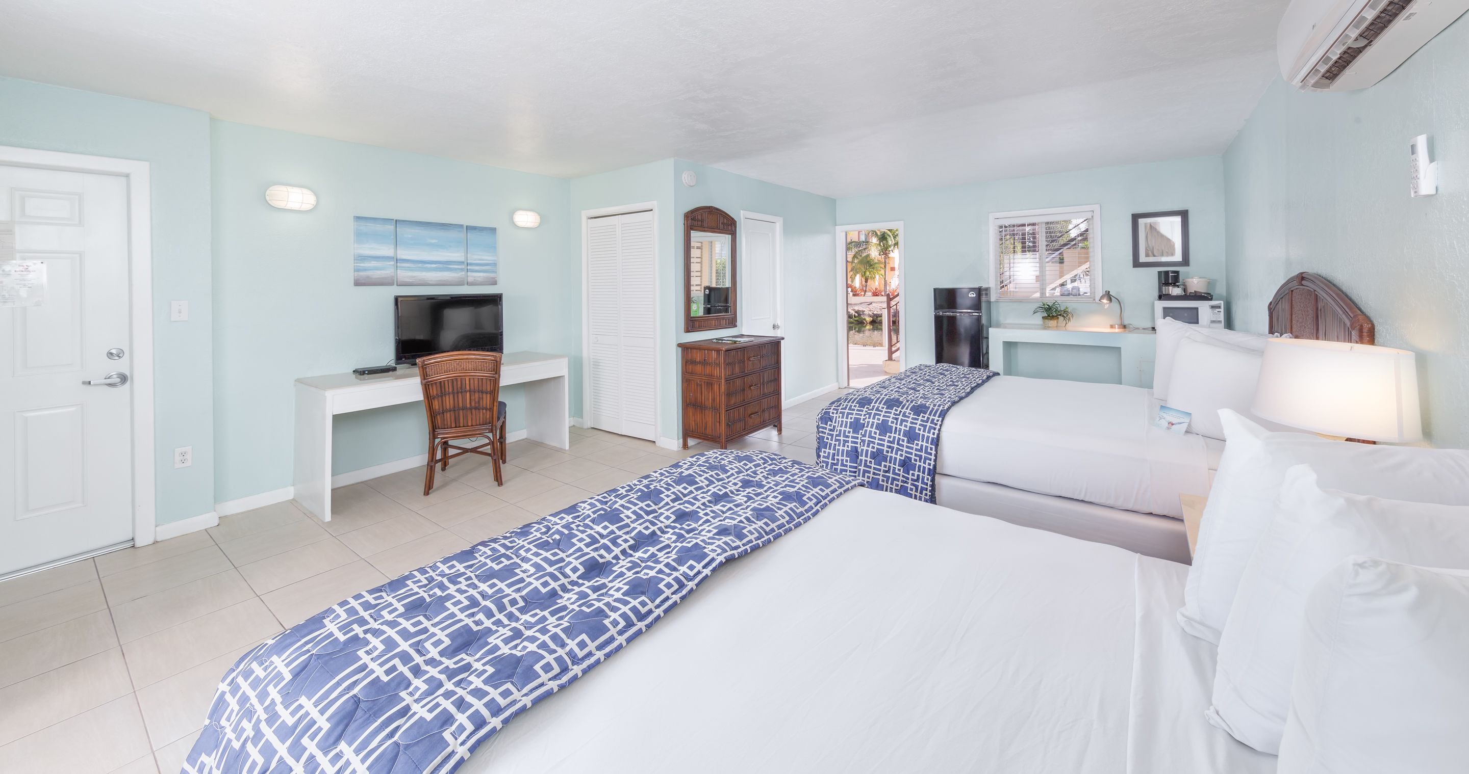 A hotel room with two beds, a desk, a chair, a television, and a window with a view of palm trees.