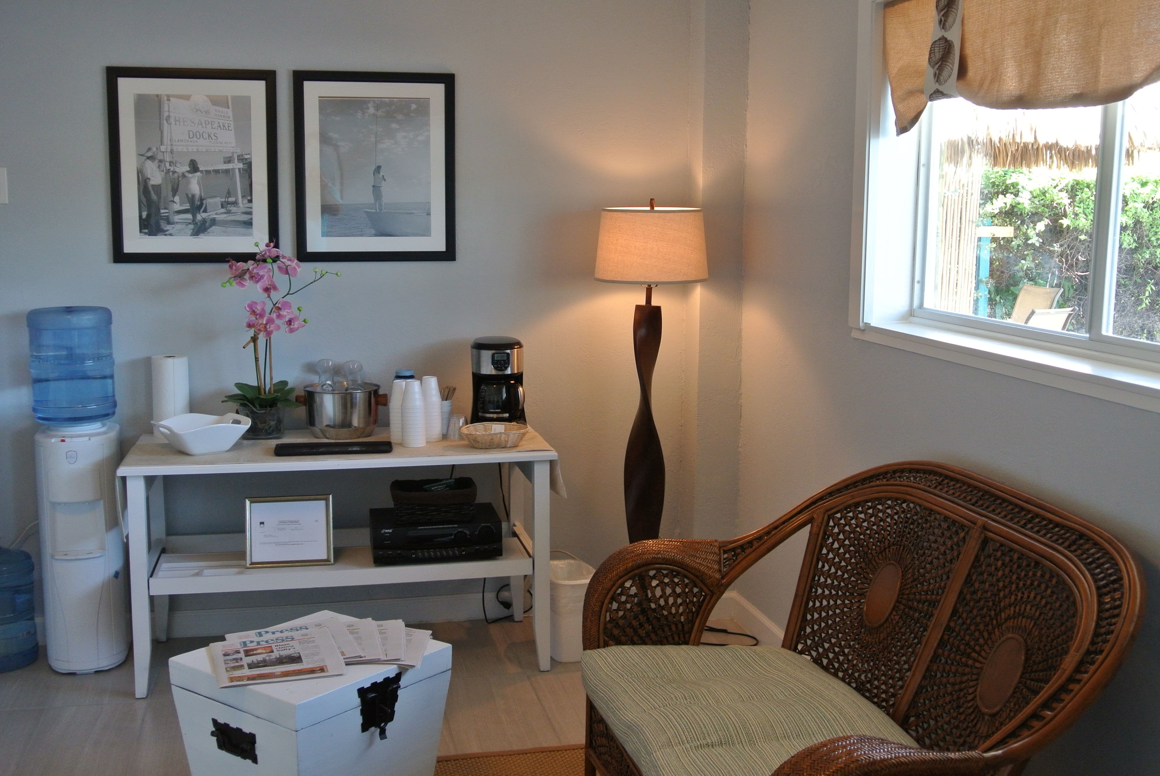 A room with a wicker chair, a desk with a lamp, framed pictures on the wall, and a water dispenser.