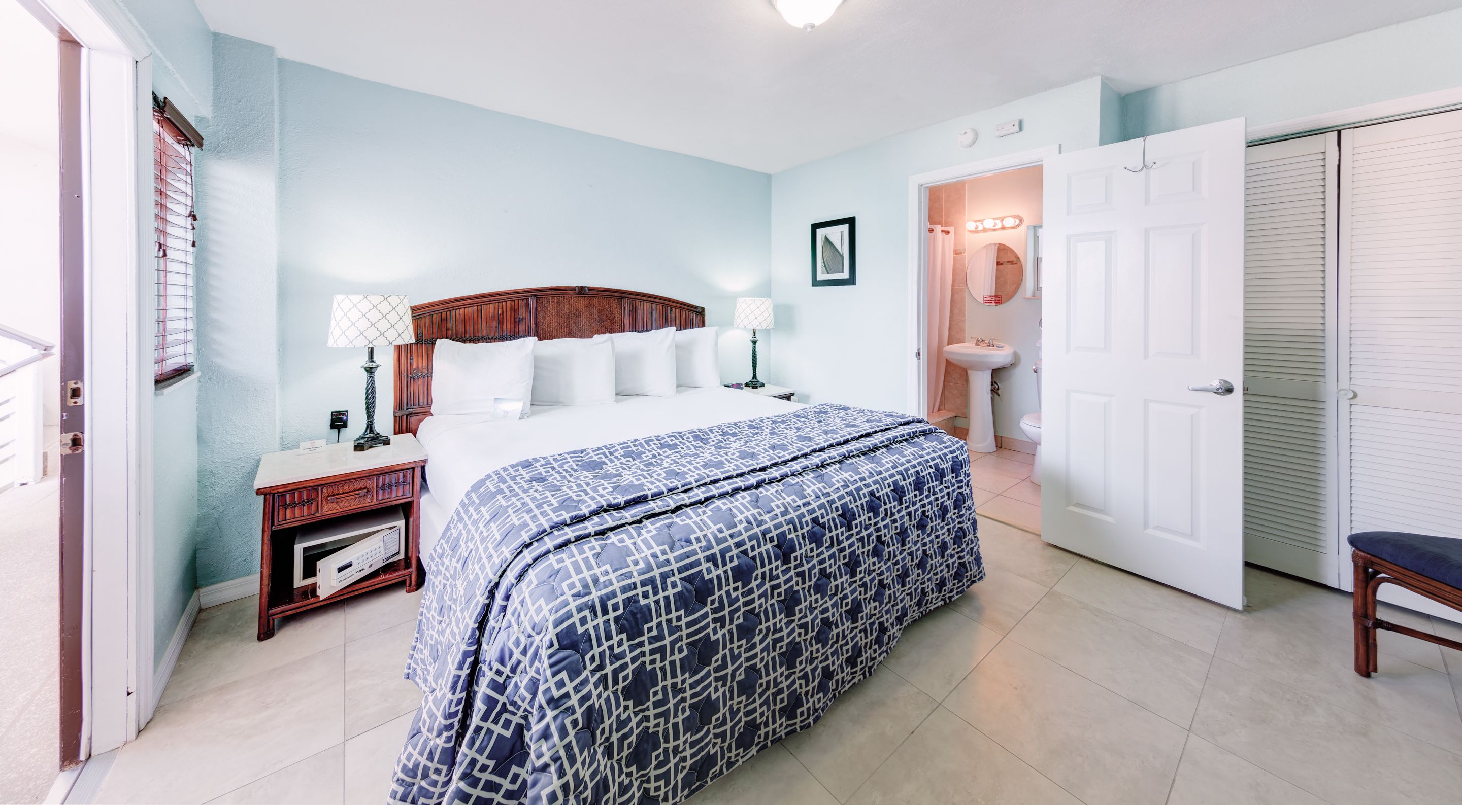 A hotel room with a bed, bedside tables, a lamp, a bathroom with a sink and mirror, and a door.