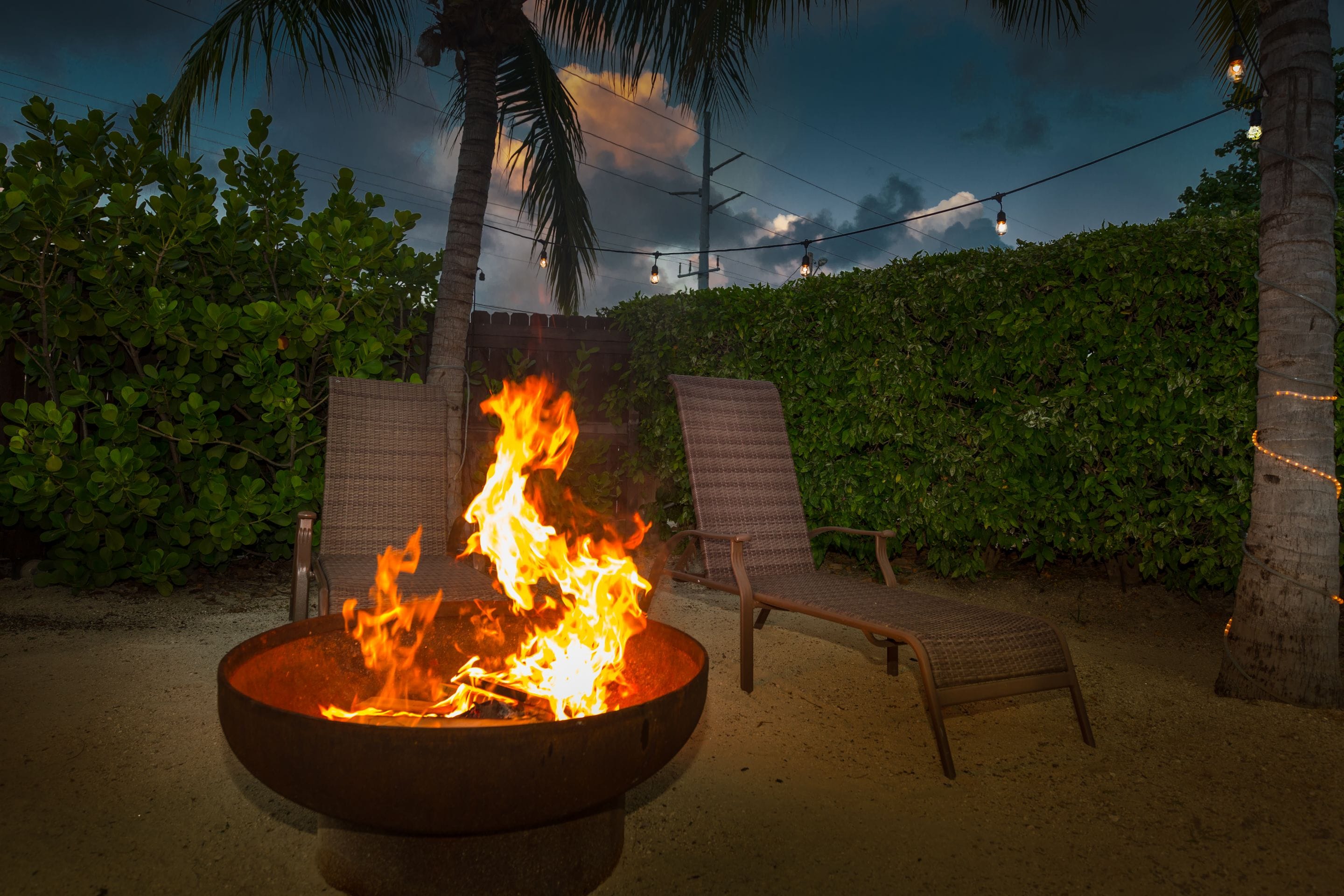 A fire pit with flames, surrounded by two wicker chairs and a palm tree.
