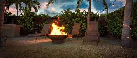 A fire pit with chairs around it, palm trees, and string lights in a backyard setting.