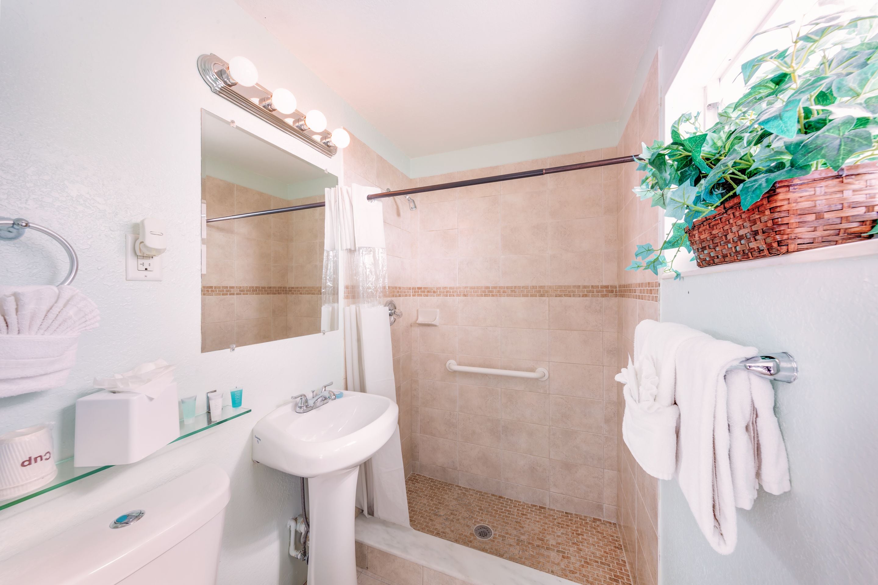 A bathroom with a glass shelf, a white sink, a mirror with lights, a shower with a handrail, and a towel rack.