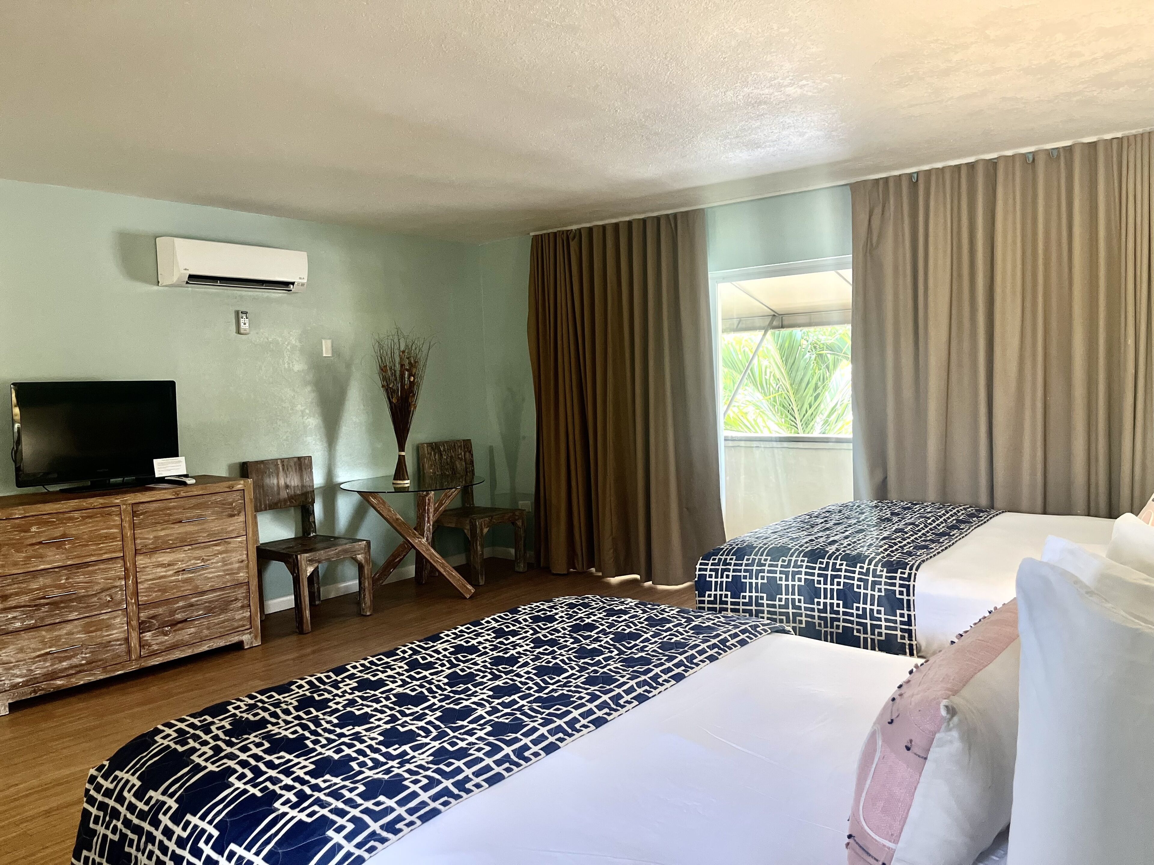 A hotel room with two beds, a wooden dresser, a television, a small table with a vase, and a window with curtains.
