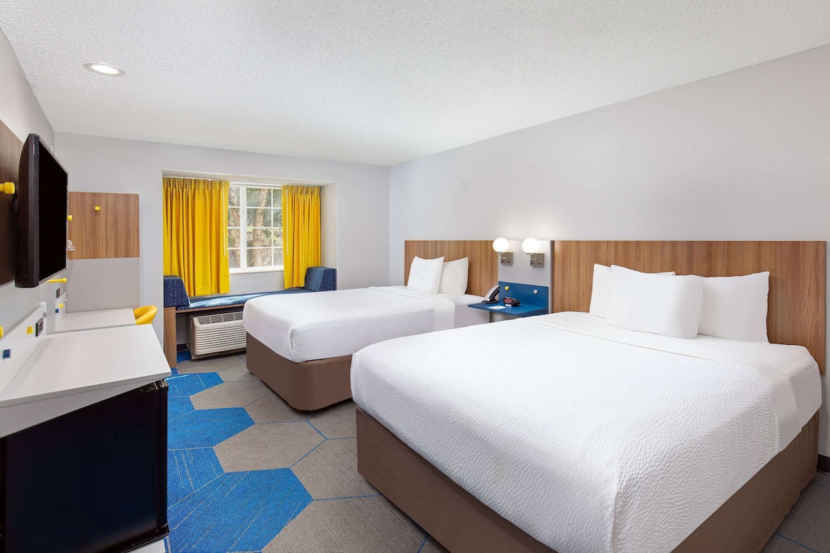 A hotel room with two beds, a desk, a chair, a TV, and a window with yellow curtains.