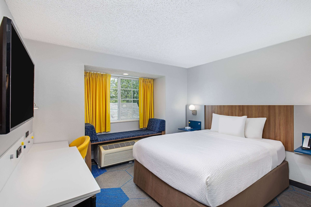 A hotel room with a large bed, a desk, a chair, a window with yellow curtains, and a television.