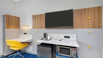A compact hotel room with a kitchenette, a small desk with a chair, a flat-screen TV, and a microwave.