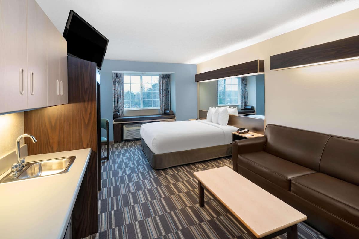 A compact hotel room with a bed, a sofa, a small table, a TV, and a kitchenette with a sink and a microwave.