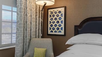 A hotel room with a bed, a chair with a patterned cushion, a lamp, and a framed artwork on the wall.