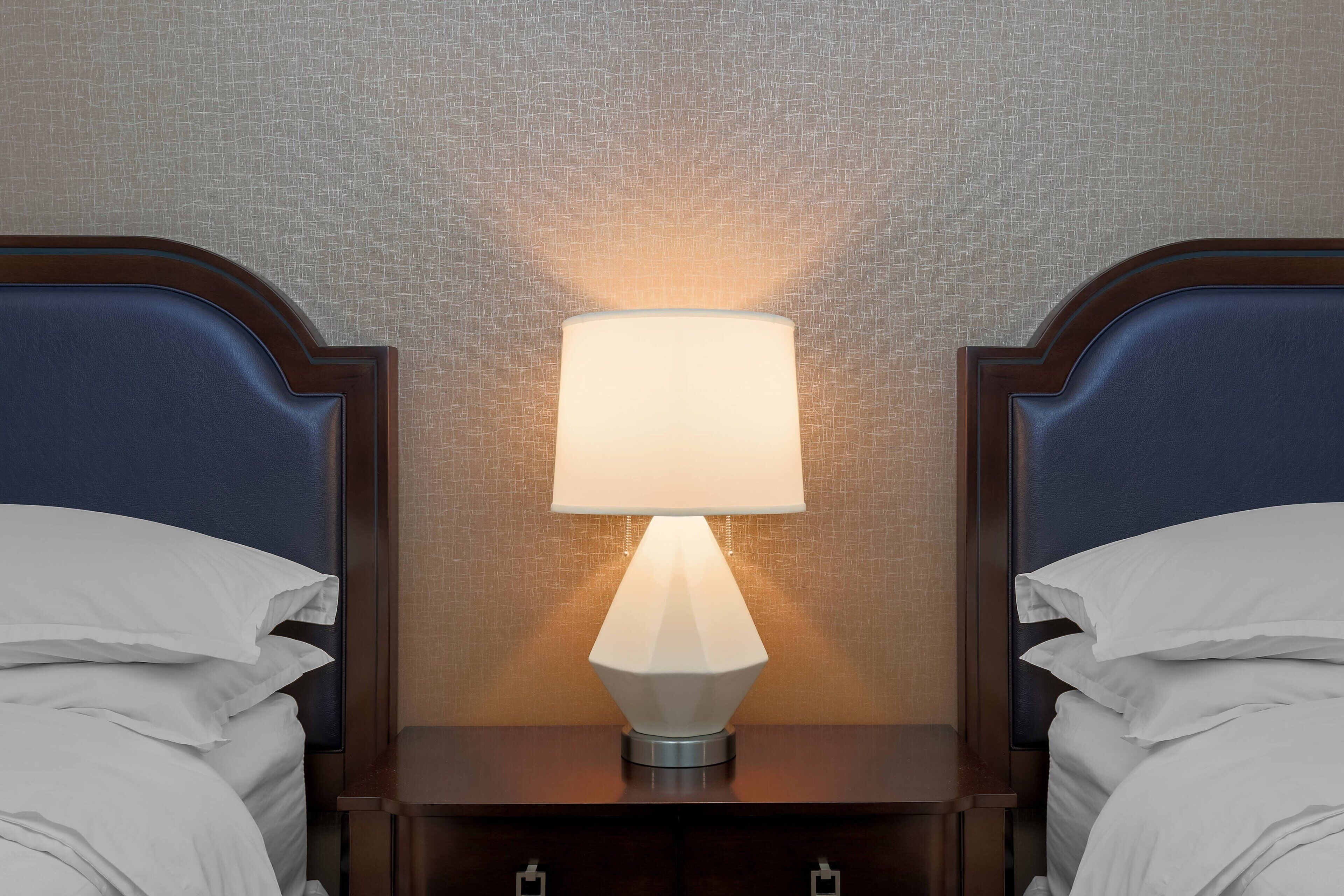 Een opgemaakt bed met witte lakens en een houten hoofdbord, een nachtkastje met een lamp, en een structuurwand op de achtergrond.