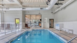 Una piscina interior con un letrero que indica una profundidad de 1,17 metros, rodeada de una zona con baldosas blancas, bancos y una puerta de cristal que da a otra habitación.