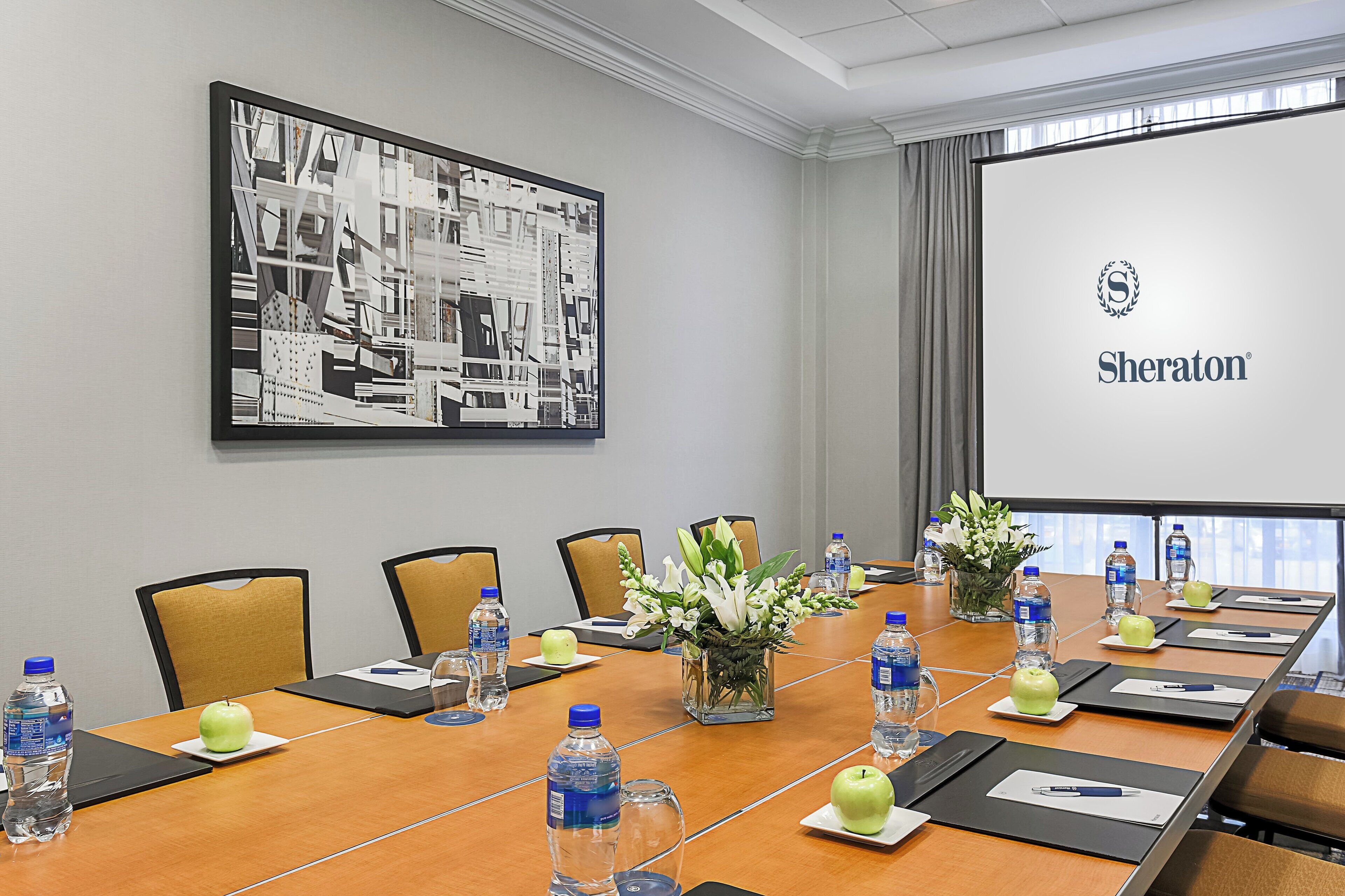 Una sala de conferencias con una larga mesa, sillas, botellas de agua, manzanas y una pantalla de proyector mostrando el logotipo de Sheraton.