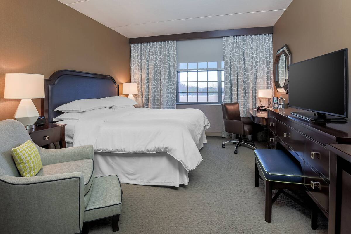 Une chambre d’hôtel avec un grand lit, un bureau équipé d’un téléviseur à écran plat, une chaise, une lampe de bureau et une fenêtre avec des rideaux.
