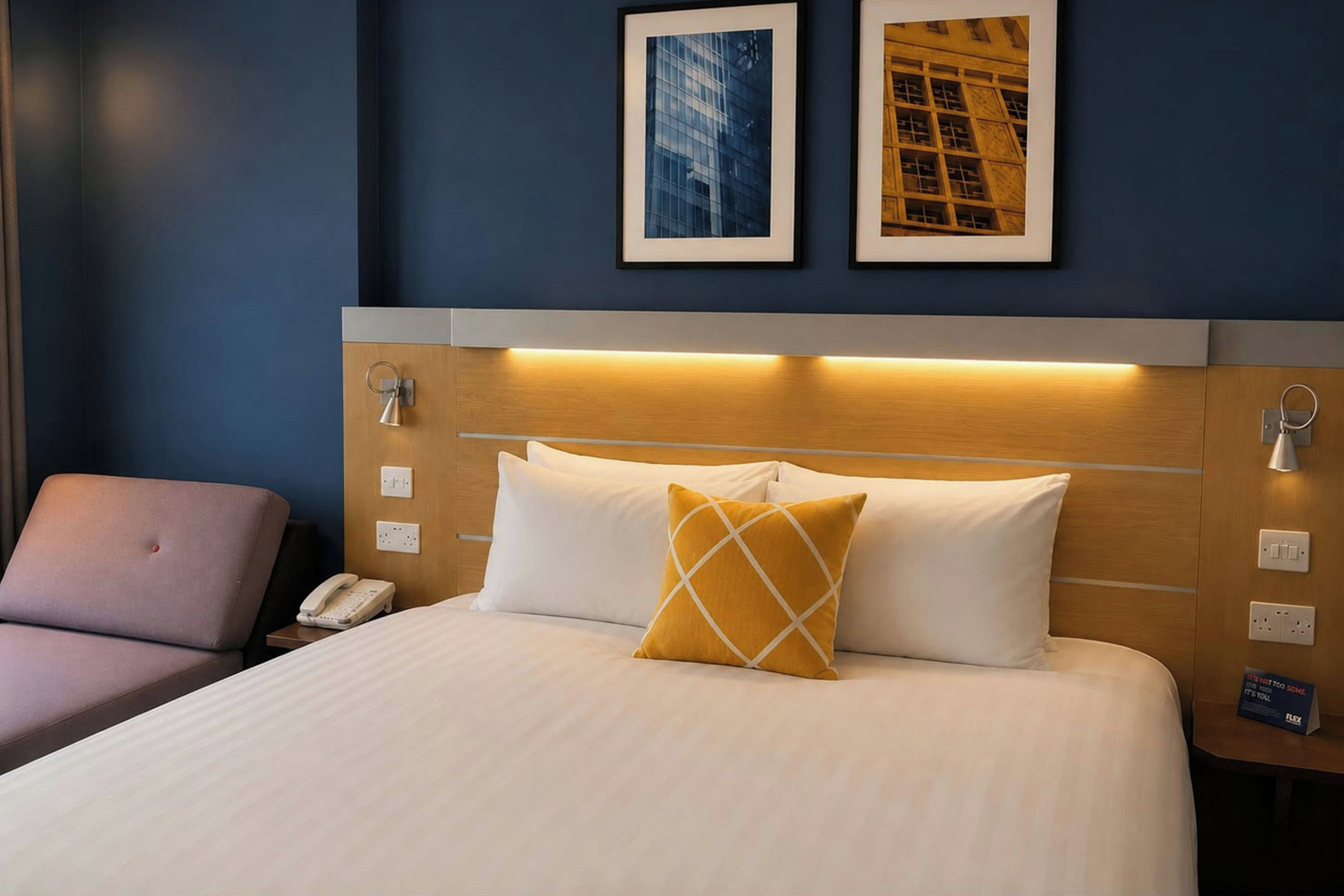 Habitación de hotel con cama, una silla, un teléfono y dos cuadros en la pared.