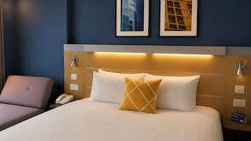 Habitación de hotel con cama, una silla, un teléfono y dos cuadros en la pared.