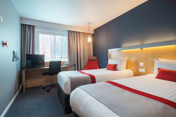Quarto de hotel com duas camas, uma escrivaninha com computador, uma cadeira e uma janela com cortinas.