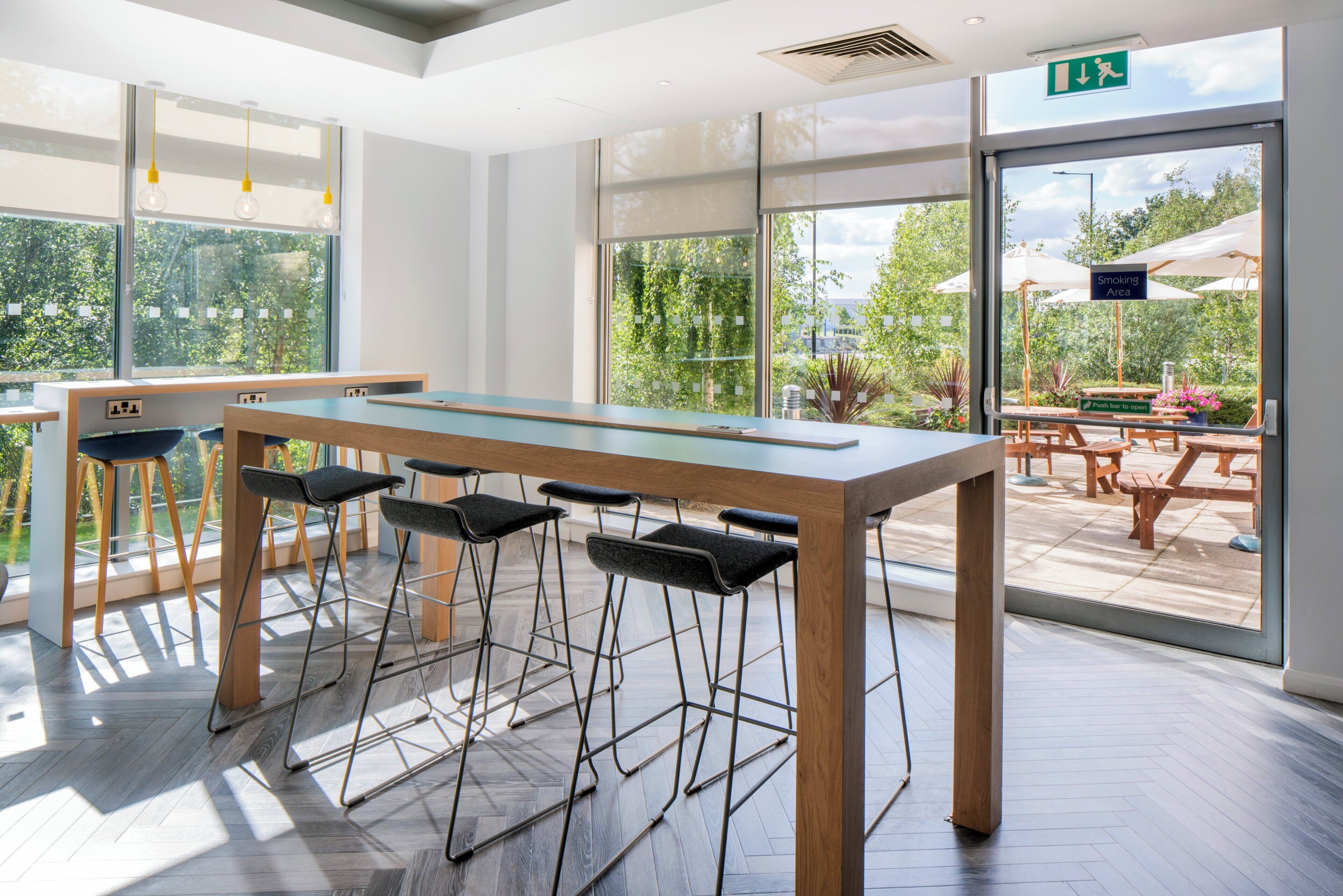 A modern indoor space with a long table and bar stools, large windows, and an outdoor patio area.