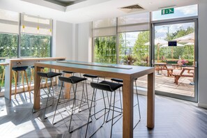 Um espaço moderno e interno com uma mesa comprida, banquetas de bar, janelas amplas e uma área de pátio ao ar livre.