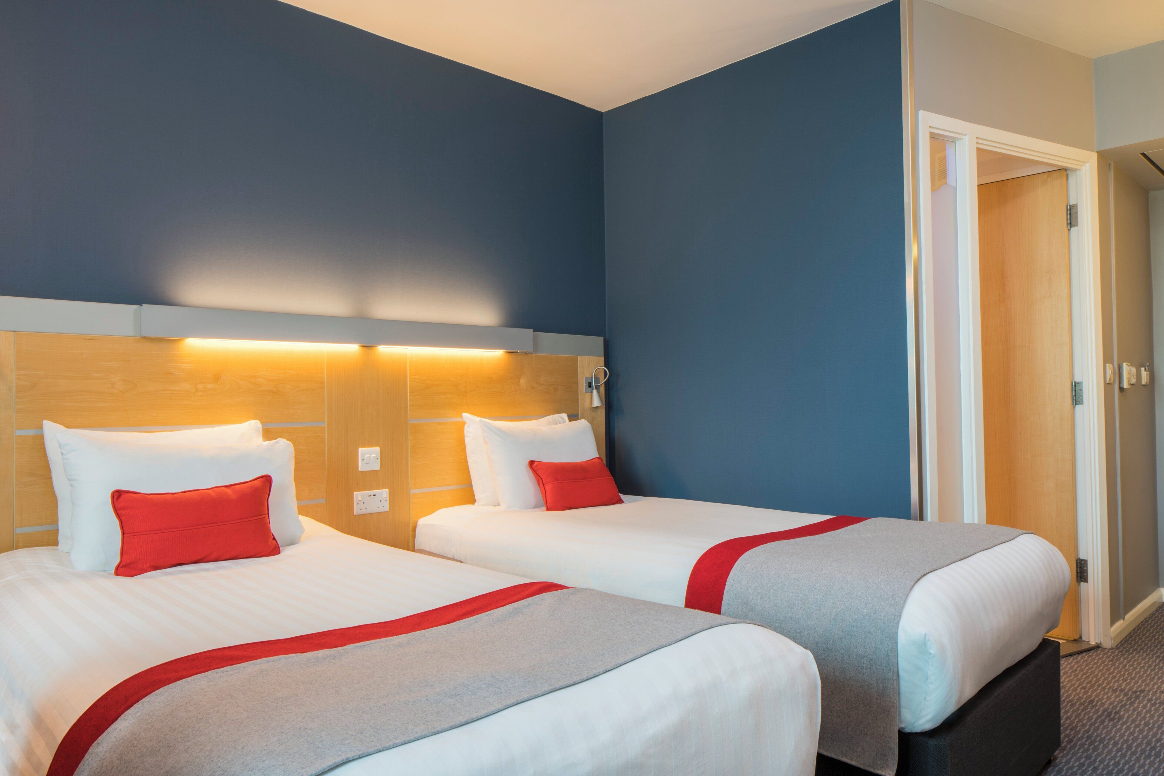 Une chambre d’hôtel avec deux lits, une tête de lit en bois, un coussin décoratif rouge, un coussin décoratif gris et un couvre-lit rouge et gris.