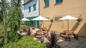 An outdoor patio with wooden tables and white umbrellas, surrounded by greenery and a brick building.