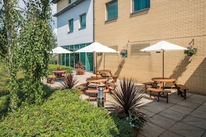 An outdoor patio with wooden tables and white umbrellas, surrounded by greenery and a brick building.