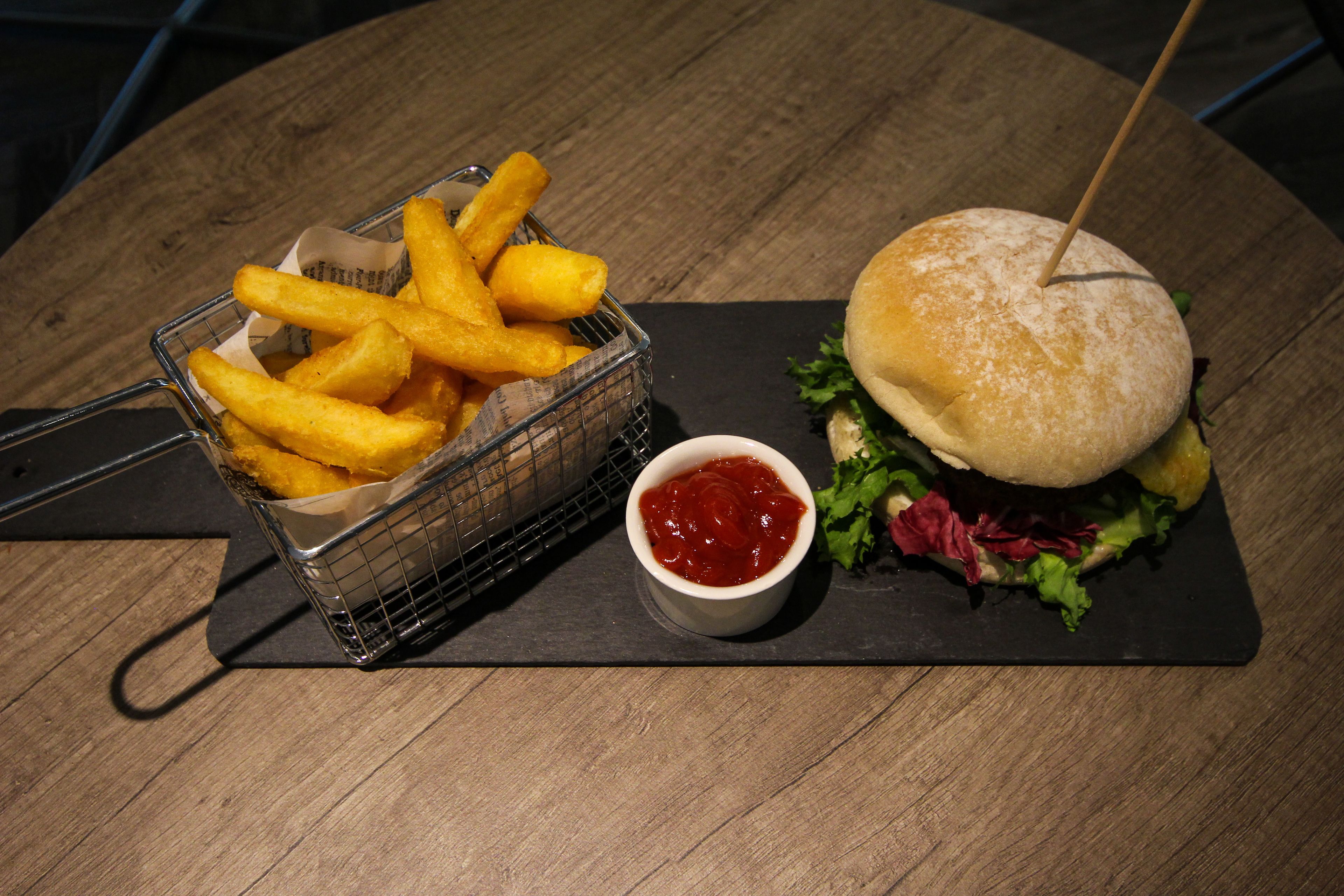 Un repas composé d’un hamburger piqué d’un cure-dent, d’un panier de frites et d’un petit bol de ketchup, le tout posé sur une table en bois.