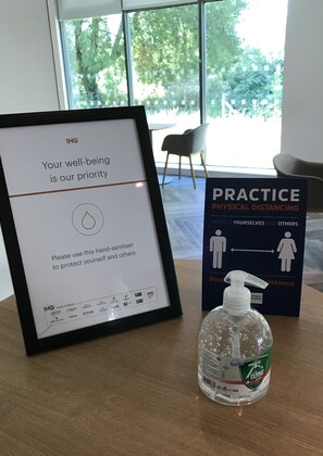 A framed sign emphasizing well-being and physical distancing, a hand sanitizer bottle, and a window view.
