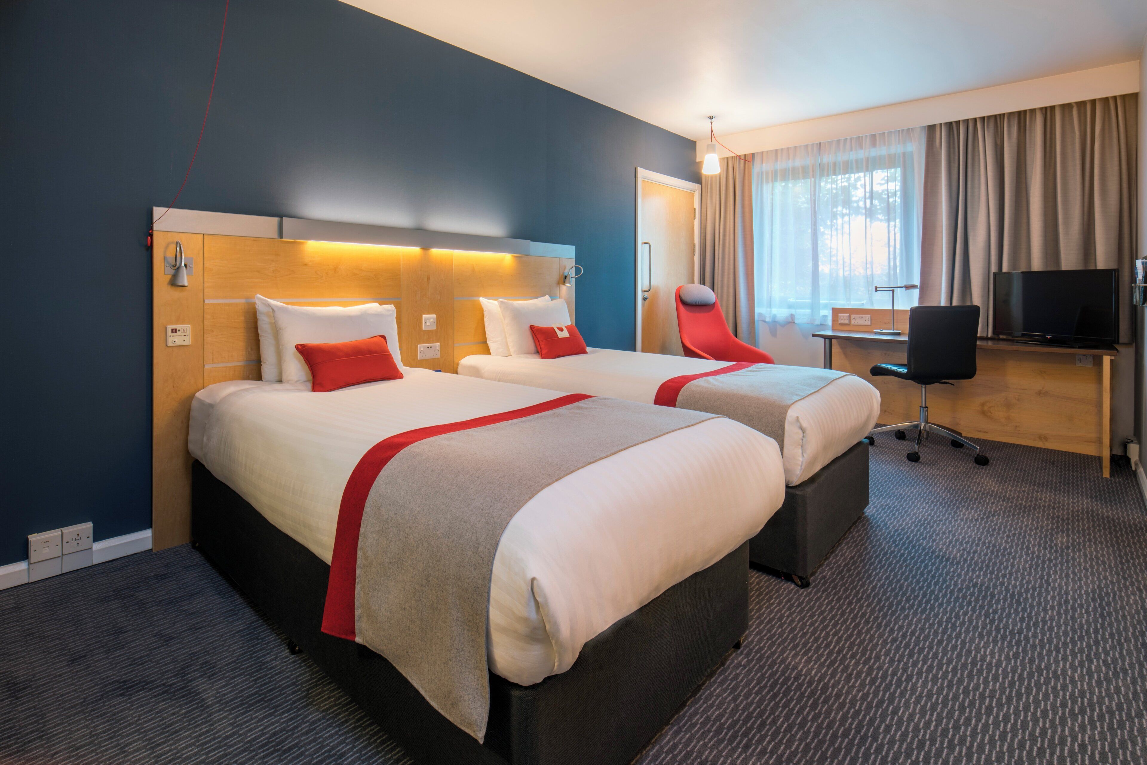 Ein Hotelzimmer mit zwei Betten, einem Schreibtisch, einem Fernseher und einem Fenster mit Vorhängen.