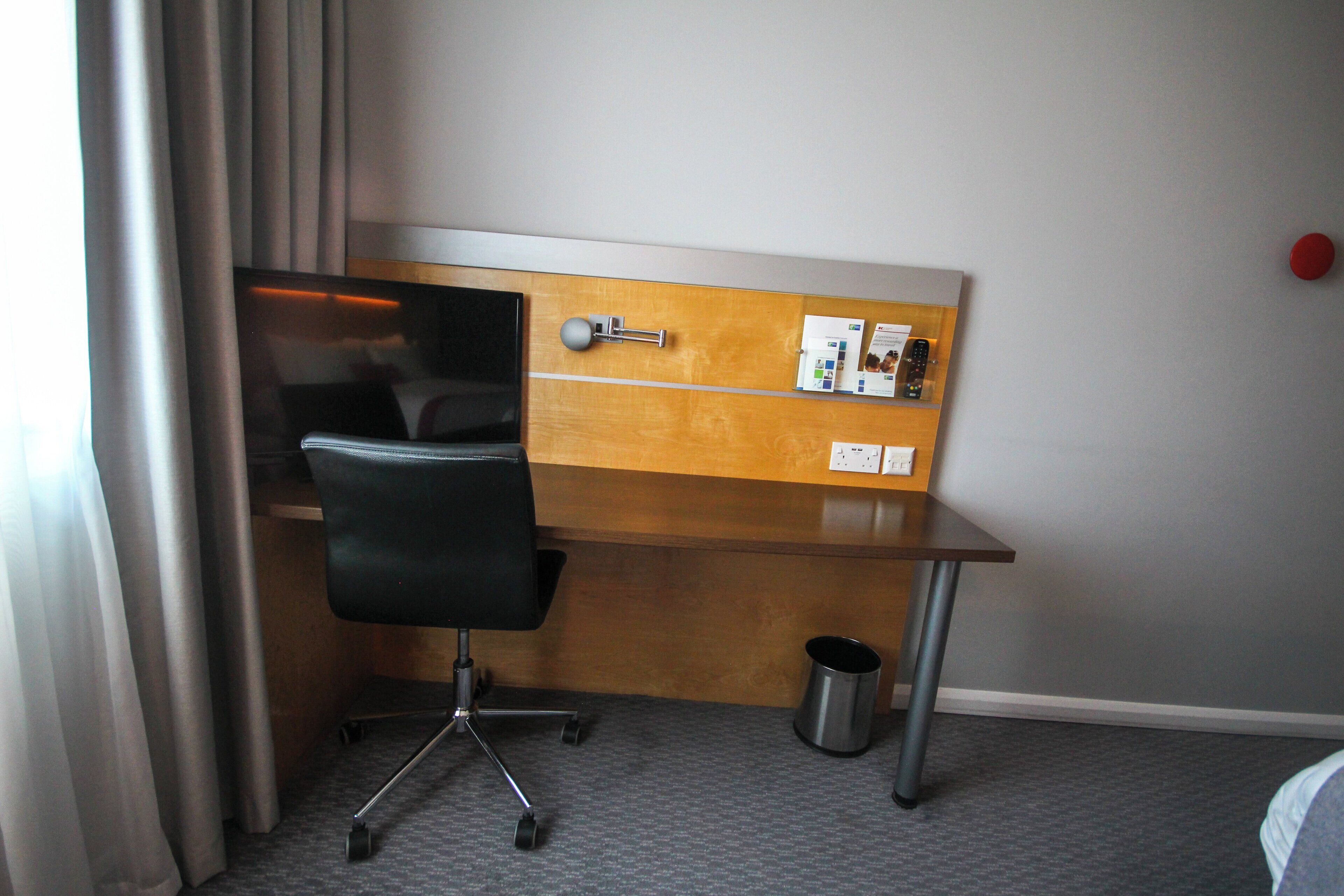 A hotel room with a desk, chair, and a small trash bin.