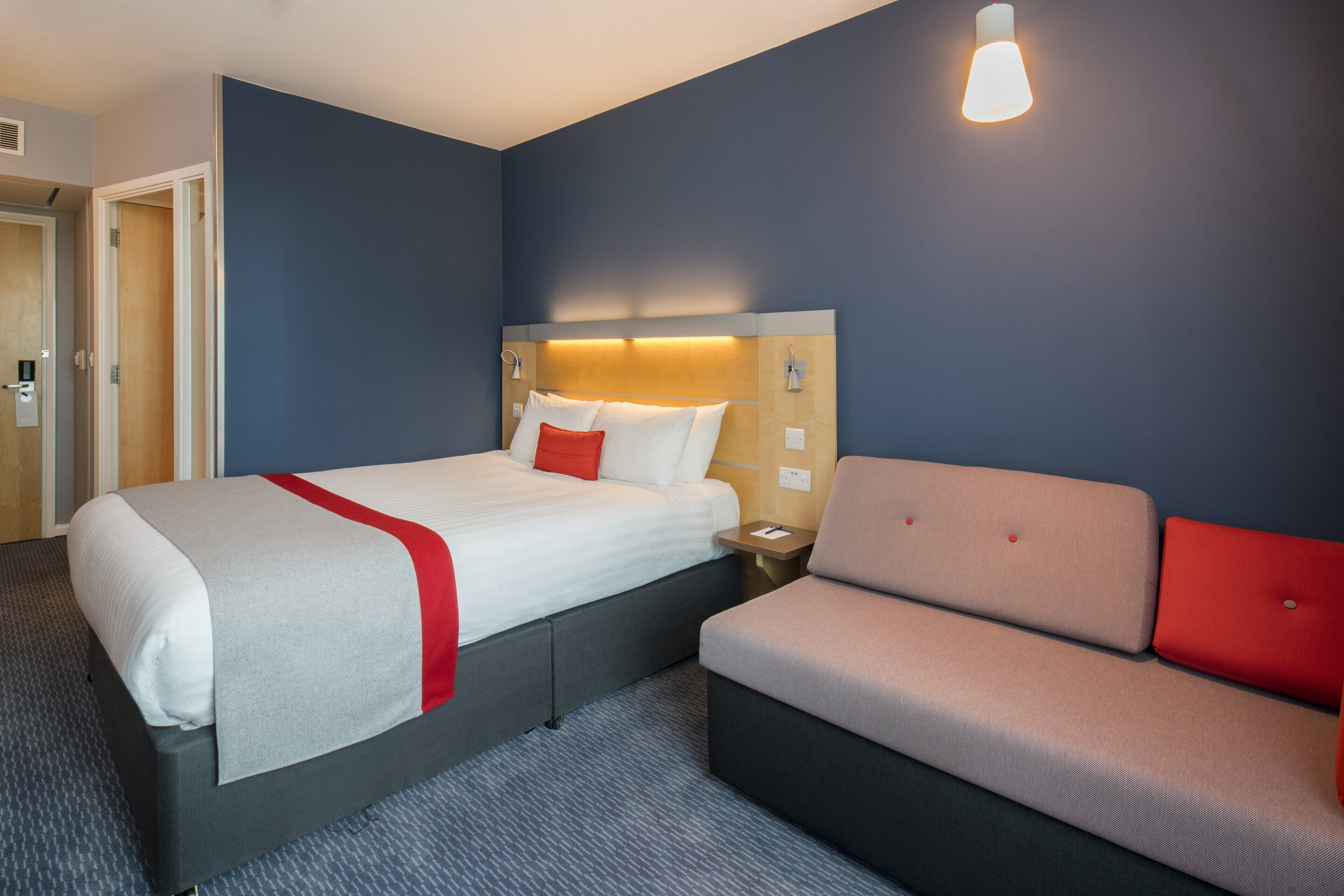 Habitación de hotel con cama, mesita de noche, sofá y lámpara de pared.