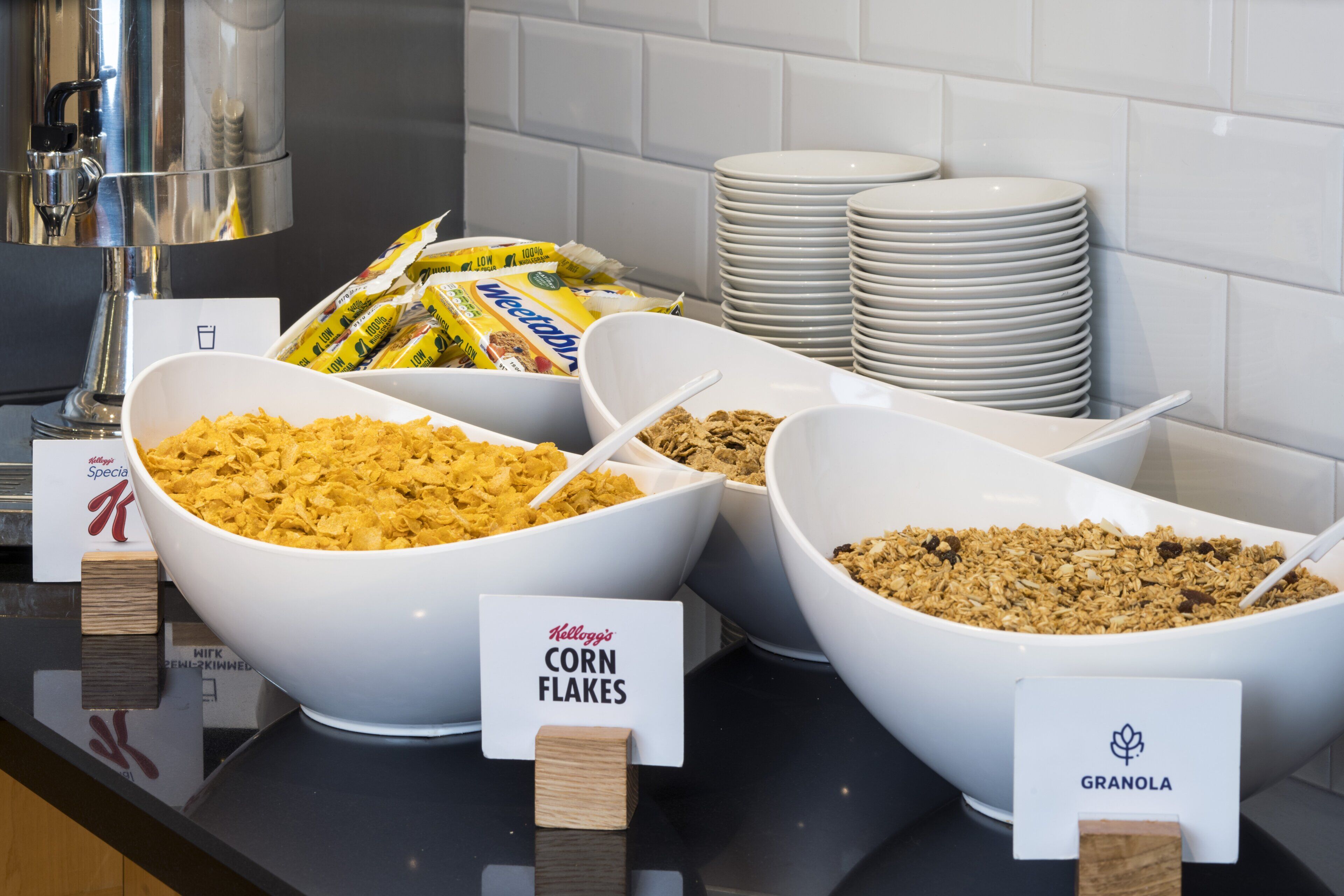 A buffet with bowls of cereal and granola, plates stacked in the background, and a coffee machine.