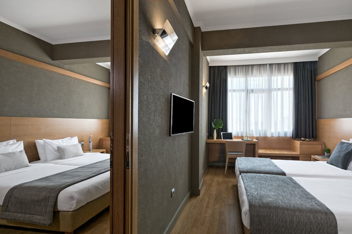 A hotel room with two beds, a desk, a TV, and a large window with curtains.